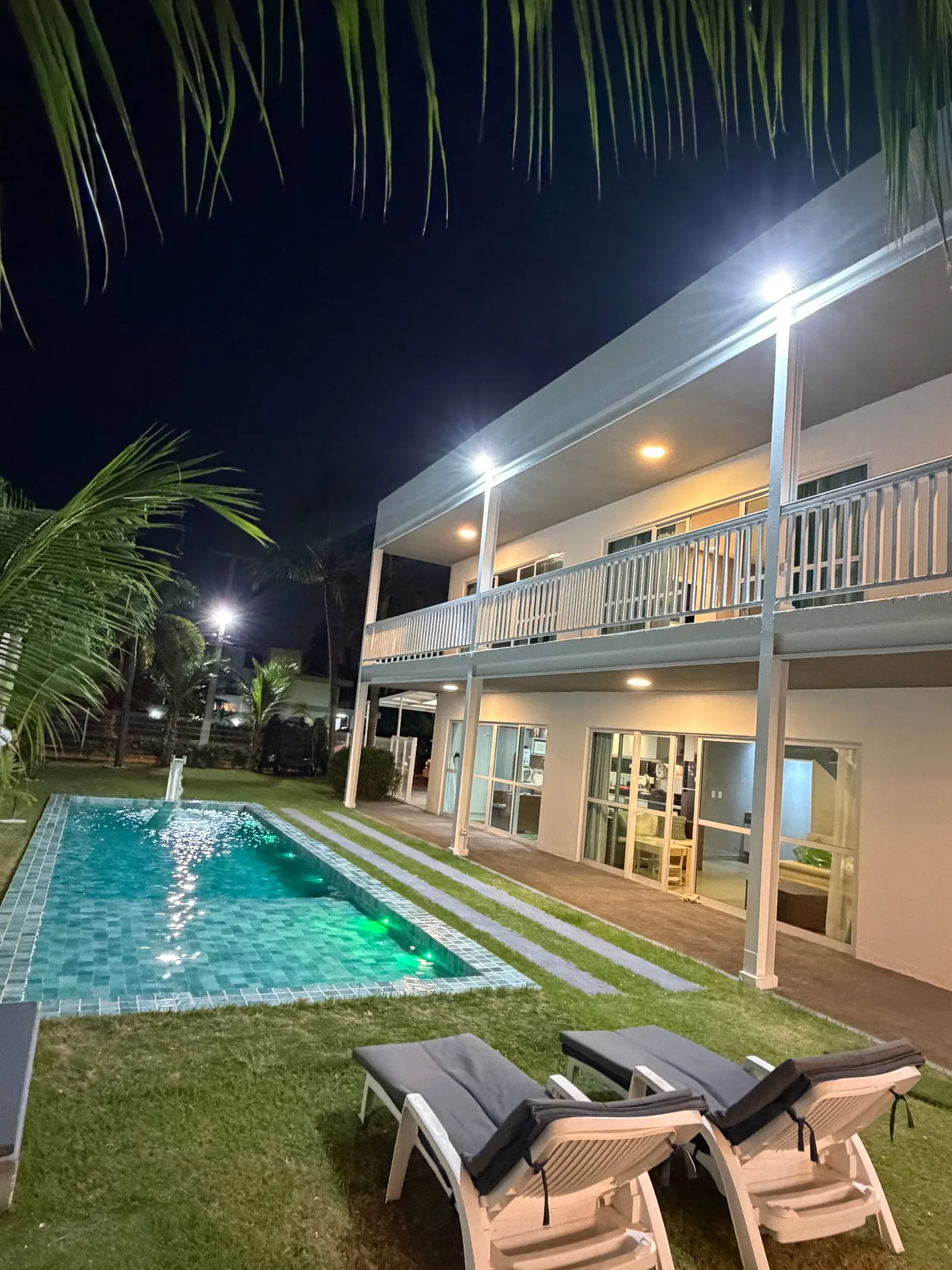 Fachada branca e piscina iluminadas à noite, coqueiros em silhueta